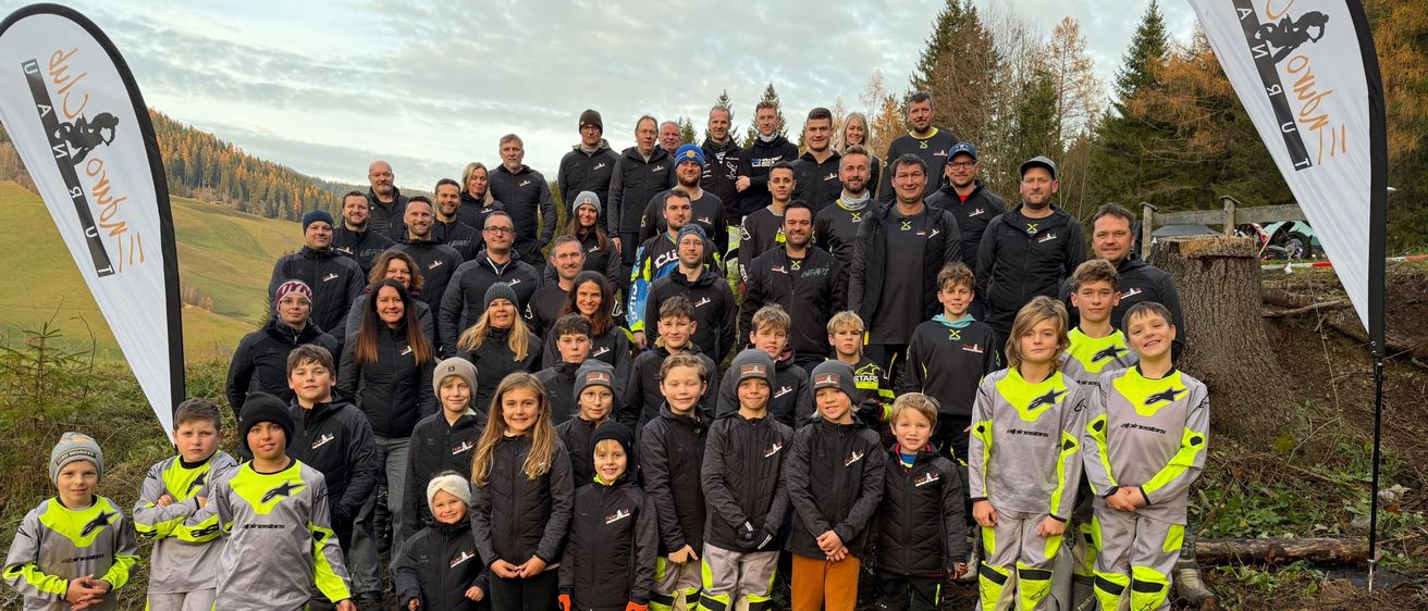 Eine Gruppenaufnahme von Erwachsenen und Kindern in Winterkleidung steht auf einem Grasfeld mit Bäumen im Hintergrund. Sie tragen passende schwarze und gelbe Jacken mit verschiedenen Logos.