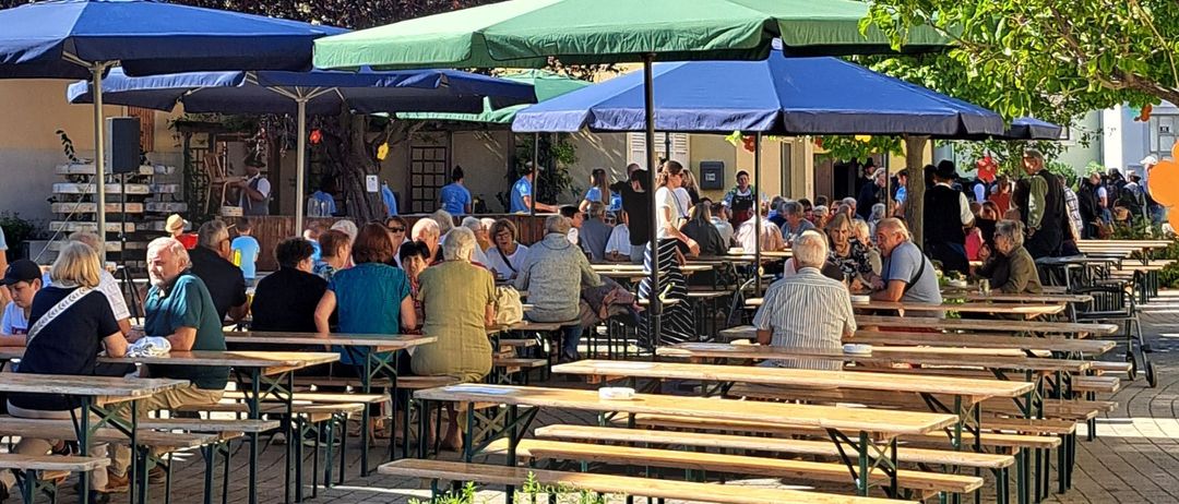 Ein sonniges Café im Freien ist mit Gästen gefüllt, die unter großen blauen und grünen Sonnenschirmen sitzen. Die Leute sitzen an Holztischen und genießen die Atmosphäre. Bäume und Gebäude sind im Hintergrund zu sehen.