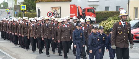 Eine Parade von uniformierten Feuerwehrleuten, einschließlich junger Kadetten, geht an einem Gebäude mit Kuppeldach und einem Feuerwehrwagen im Hintergrund vorbei.