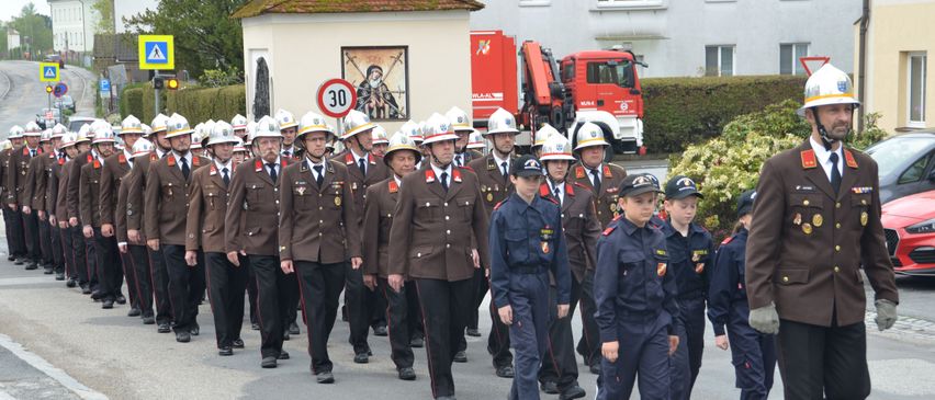 A group of firefighters in uniform, some with hats, walk in a line. Behind them is a building with a red sign and a truck.