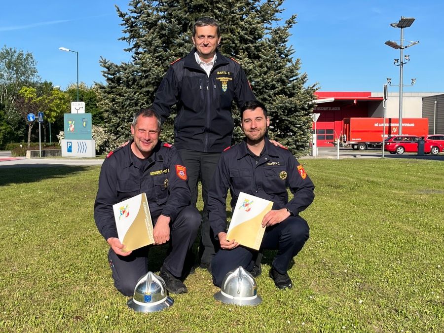 Drei Feuerwehrleute posieren für ein Foto mit Urkunden und Helmen vor einer Feuerwache, wobei ein roter LKW und ein Auto dahinter geparkt sind.