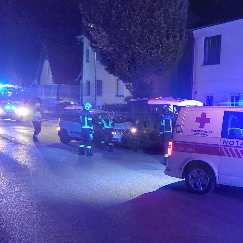 Ein Rettungswagen und ein Feuerwehrwagen stehen nachts auf einer Straße. Zwei Personen in Uniform stehen neben einem Auto.
