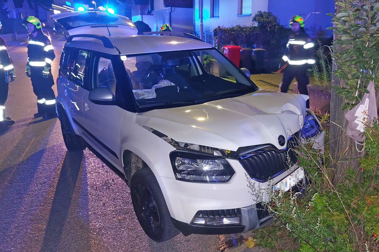 Ein weißer SUV mit beschädigter Front ist am Straßenrand geparkt. Ein Feuerwehrmann in Uniform steht in der Nähe, und eine weitere Person sitzt im Fahrzeug.