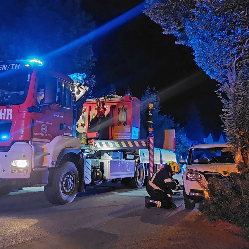 Ein Feuerwehrauto steht nachts auf der Straße. Ein Feuerwehrmann kniet und arbeitet an einem Auto. Ein anderer Feuerwehrmann ist auf dem LKW.