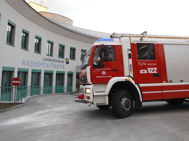 Ein rot-weißer Feuerwehrwagen fährt vor einem Gebäude vorbei, das mit Landeklinikum Waidhofen/Thaya beschriftet ist.