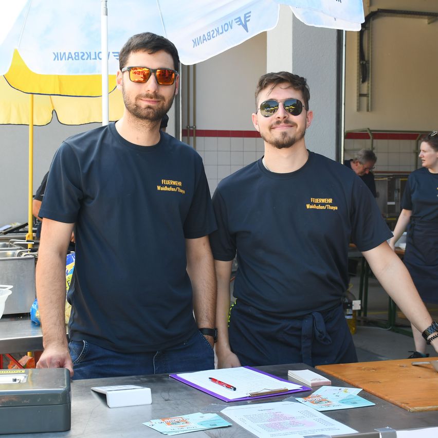Zwei Männer in blauen T-Shirts stehen hinter einem Tisch und tragen eine Sonnenbrille. Die T-Shirts haben gelben Text und sie befinden sich in einem Raum mit einem Regenschirm und einer Wand mit Fliesen.