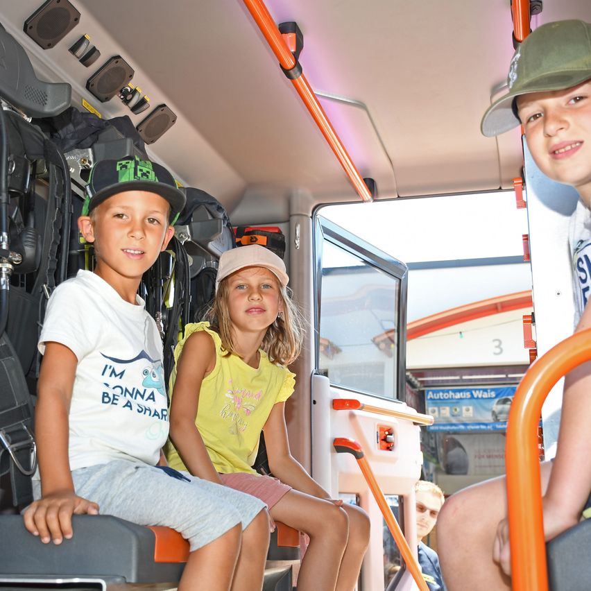 Drei Kinder sitzen in einem Krankenwagen, zwei vorne und eines an der Seite. Der Bus hat orangefarbene Handläufe, und ein Schild, das 'Autohaus Wais' anzeigt, ist draußen sichtbar.