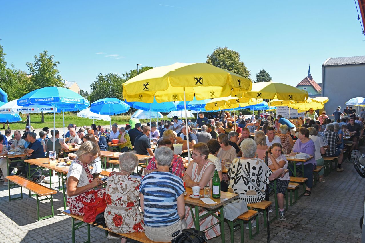 Eine Menschenmenge sitzt an Picknicktischen im Freien. Die Tische sind mit blauen und gelben Sonnenschirmen bedeckt. Die Menschen genießen Essen und Getränke. Der Himmel ist blau mit einigen Wolken.