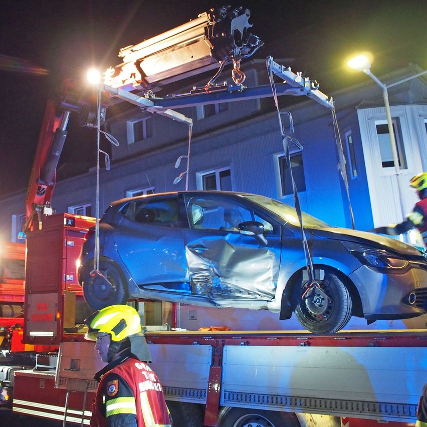 Ein blaues Auto wird nachts von einem Kran angehoben und zeigt schwere Schäden an der Front. Zwei Feuerwehrleute in gelben Helmen sind vor Ort. Ein Feuerwehrwagen und ein Gebäude sind im Hintergrund zu sehen.