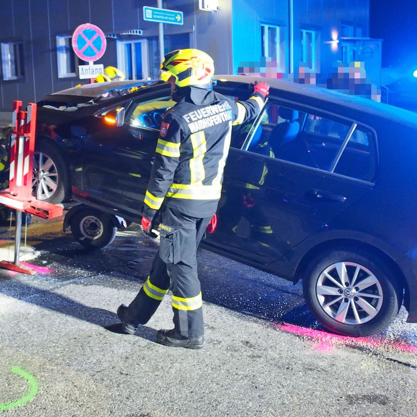 Ein Feuerwehrmann hält ein demoliertes Auto, das von einem Wagenheber angehoben wird, während zwei andere im Inneren sind. Die Szene spielt sich draußen bei Nacht ab.