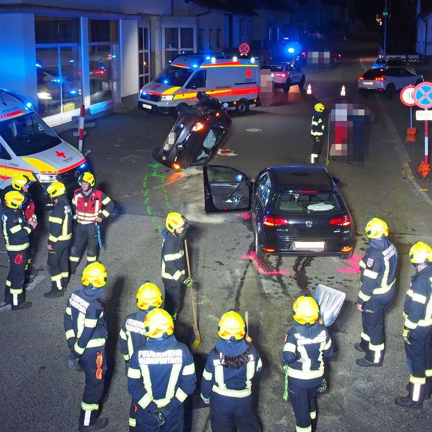 Rettungskräfte stehen um ein umgekipptes Auto und ein Auto mit offener Tür herum. Es gibt Rettungsfahrzeuge und Verkehrskegel um die Szene.