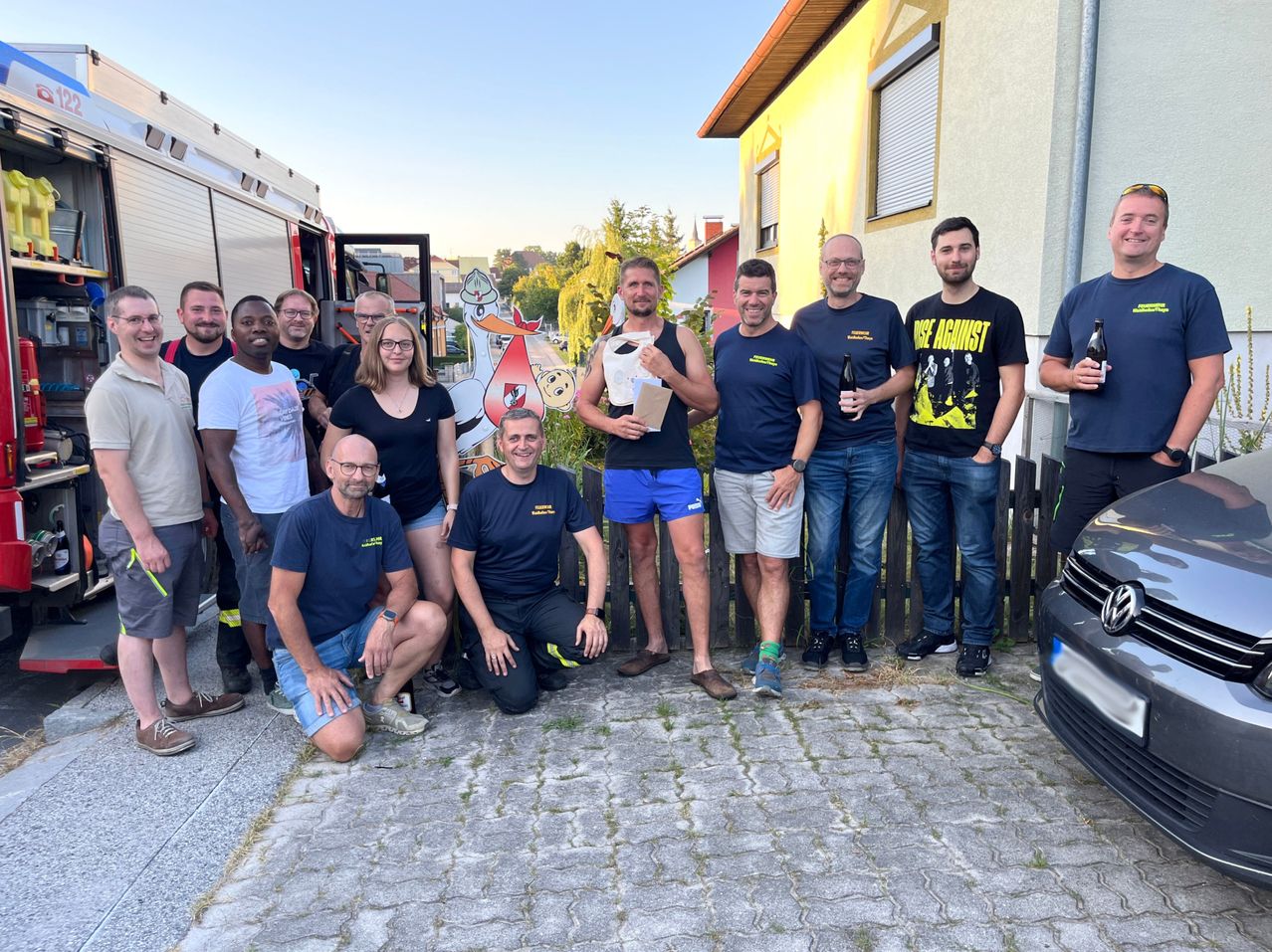 Eine Gruppe Feuerwehrleute posiert für ein Foto vor einem Haus, einer hält eine Box, der andere eine Flasche.