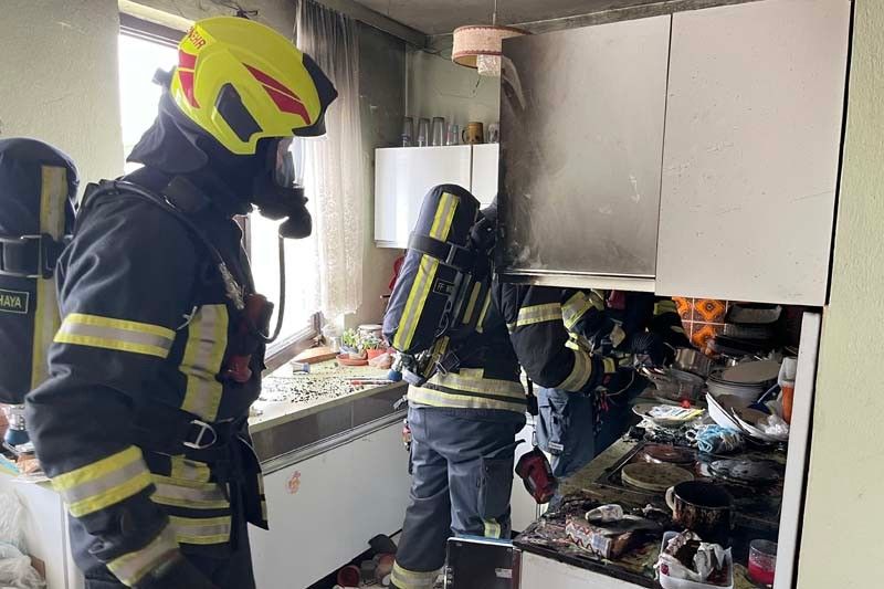 Drei Feuerwehrleute in Schutzkleidung sind in einer Küche. Die Küche hat einen gefliesten Boden, weiße Schränke und eine beschädigte Arbeitsplatte. Der Bereich sieht rauchig aus.