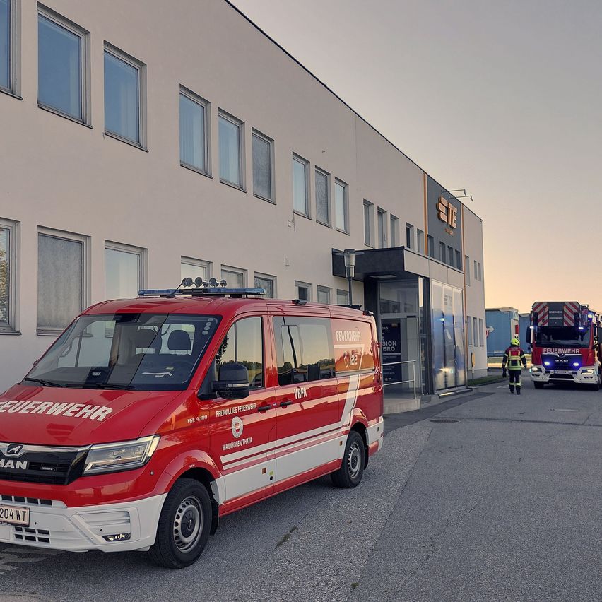 Ein roter Feuerwehrwagen ist vor einem Gebäude mit mehreren Fenstern geparkt. Ein Feuerwehrwagen ist hinter dem Van geparkt. Eine Person in einer Sicherheitsjacke steht in der Nähe des Feuerwehrwagens.