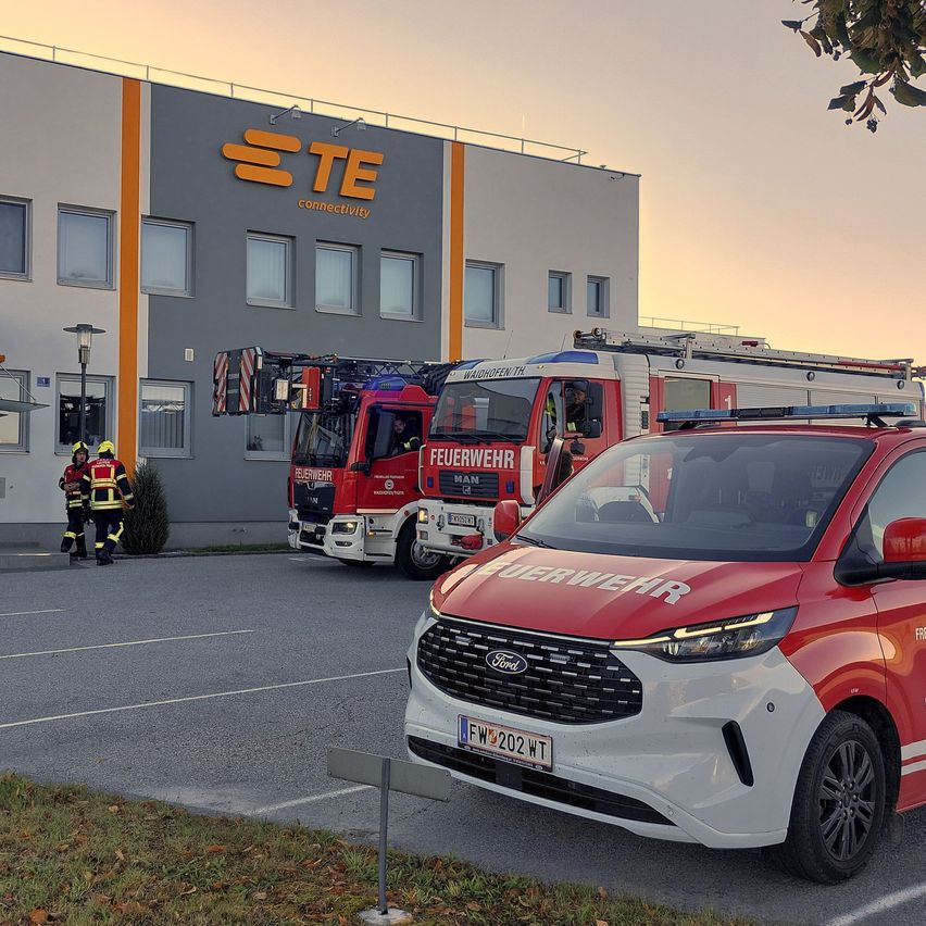 Ein Feuerwehrwagen mit der Aufschrift 'Feuerwehr' steht vor einem Gebäude mit 'TE Connectivity' an der Seite. Zwei Feuerwehrleute stehen neben dem Gebäude.