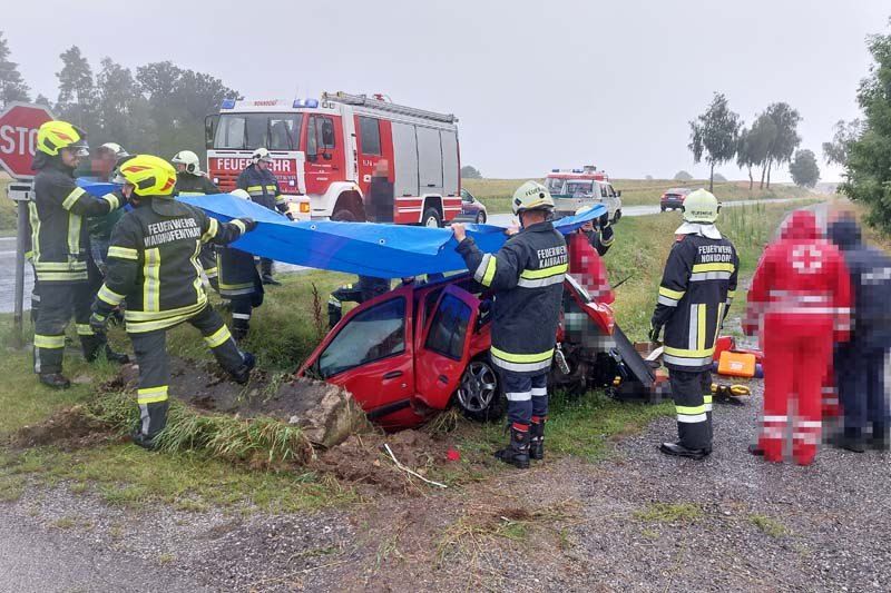 Feuerwehrleute entfernen ein rotes Auto von der Straße nach einem Unfall. Rettungsfahrzeuge sind in der Nähe geparkt.