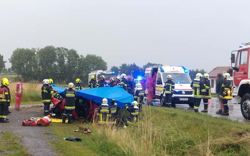Rettungskräfte kümmern sich um ein beschädigtes Fahrzeug mit einer blauen Abdeckung. Sie tragen Helme und Uniformen. Ausrüstung und ein Feuerwehrwagen sind in der Nähe.
