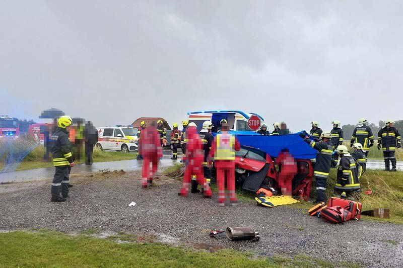 Einsatzkräfte in Schutzkleidung umgeben ein beschädigtes rotes Auto am Straßenrand, mit einem Stoppschild im Hintergrund.