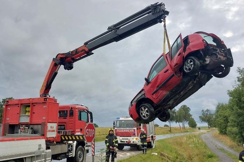 Ein rotes Auto wird von einem Kran gehoben, möglicherweise nach einem Unfall geborgen. Zwei Feuerwehrleute beobachten vom Straßenrand aus.