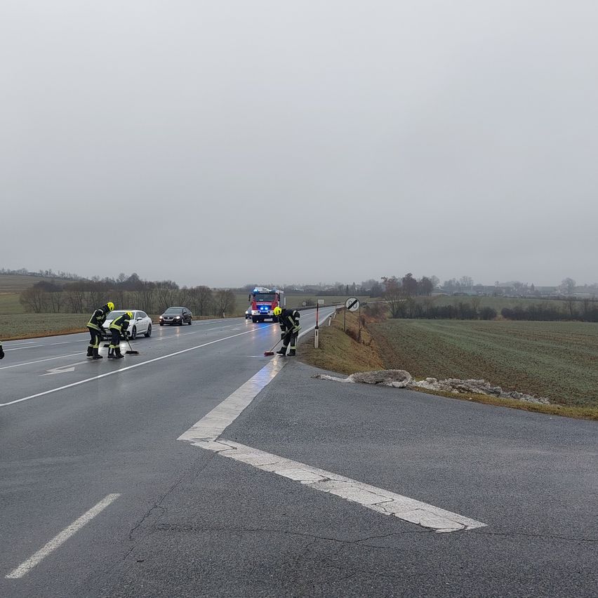Rettungskräfte reinigen eine Straße. Zwei Personen benutzen Besen und Kehrschaufel. Ein Feuerwehrauto ist am Straßenrand geparkt.