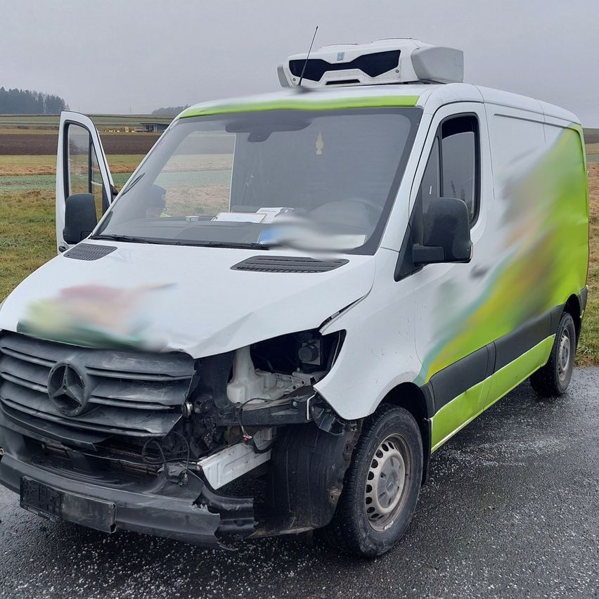 Ein beschädigter Mercedes-Benz Sprinter-Transporter ist auf einer Straße geparkt, wobei die Front stark beschädigt und die Tür offen ist.