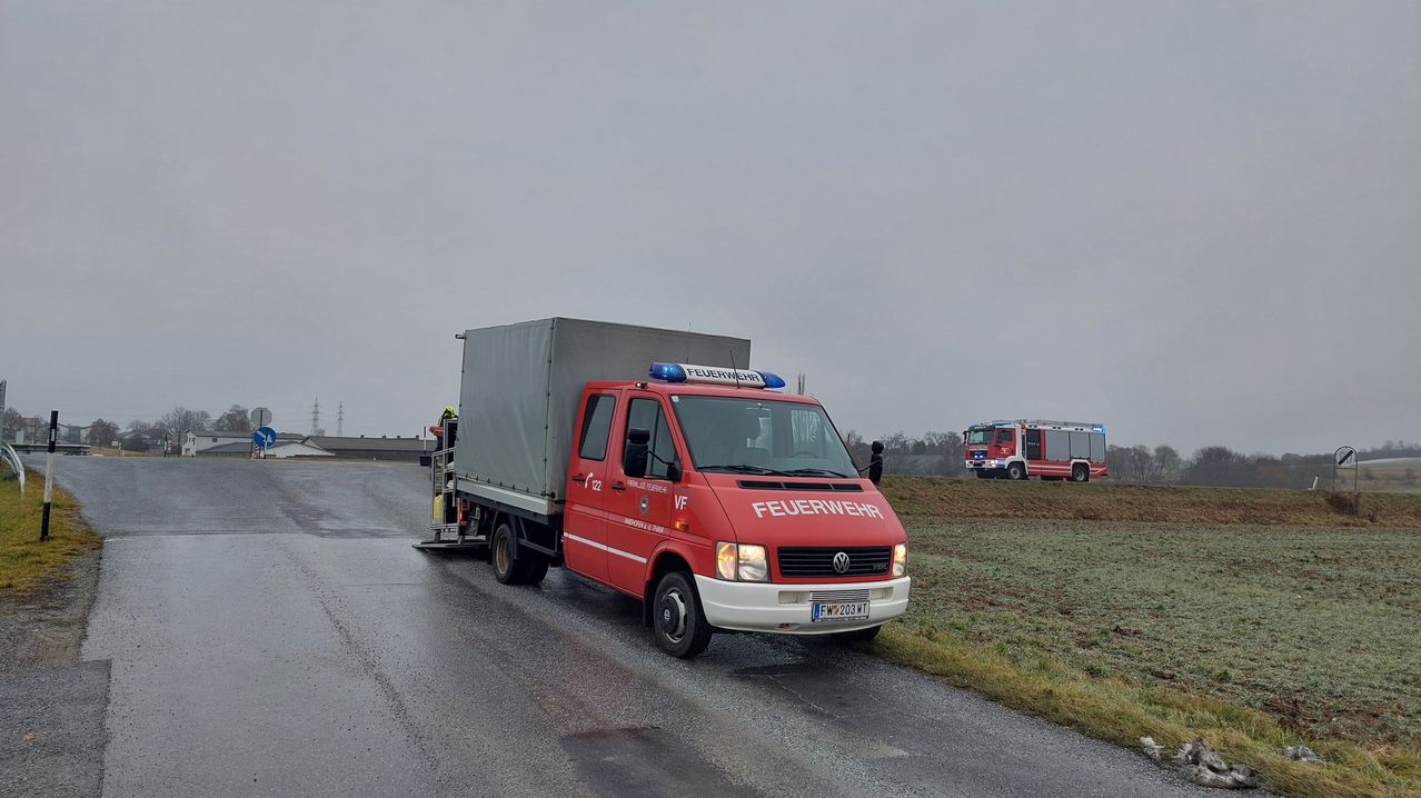 Ein roter Feuerwehrwagen steht auf einer nassen Straße, mit einem weiteren Feuerwehrwagen im Hintergrund auf einem grasigen Feld unter einem bewölkten Himmel.