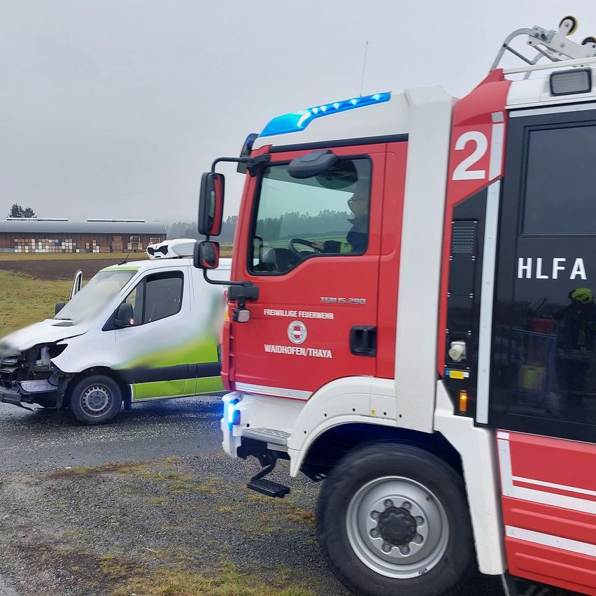 Ein roter Feuerwehrwagen mit der Nummer 21 steht am Straßenrand. Ein weißer Lieferwagen ist davor geparkt und hat vorne schwere Schäden erlitten.
