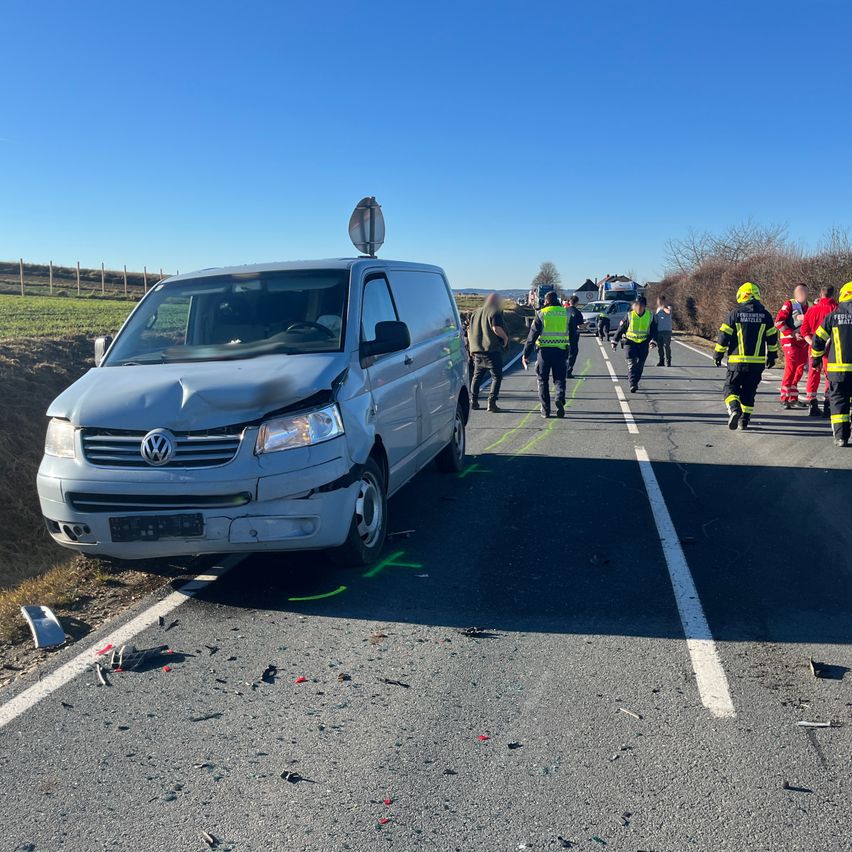 A Volkswagen van, damaged in a collision, is parked on the roadside with emergency services attending. People in safety vests are walking towards the van.
