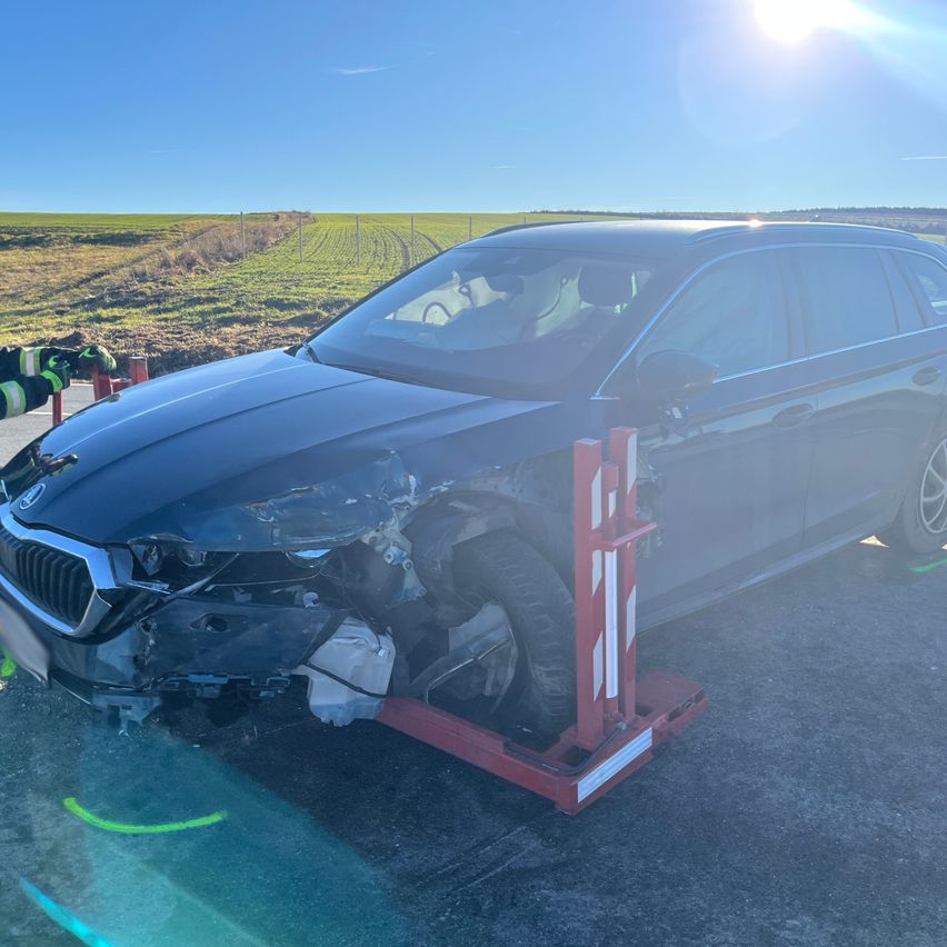 A black BMW car with a damaged front is being lifted by a red tow truck on a sunny day. The car has a flat tire on the back.