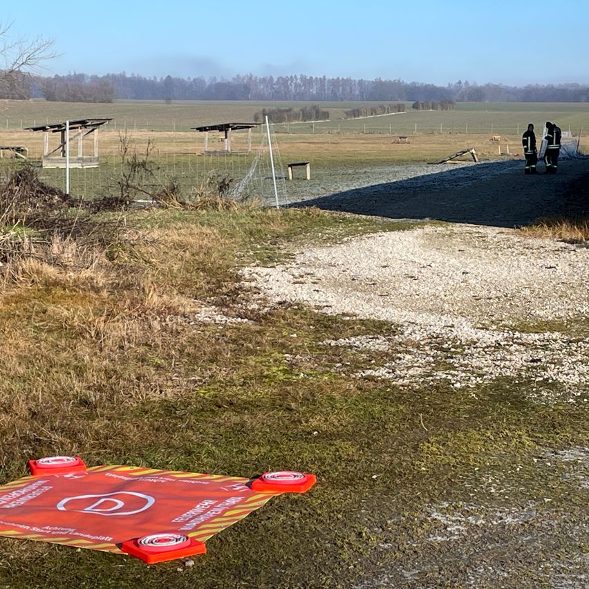 Ein offenes Feld mit zwei Personen in Schutzkleidung, die unter einer Struktur stehen. Eine rote Sicherheitsmatte liegt auf dem Boden.