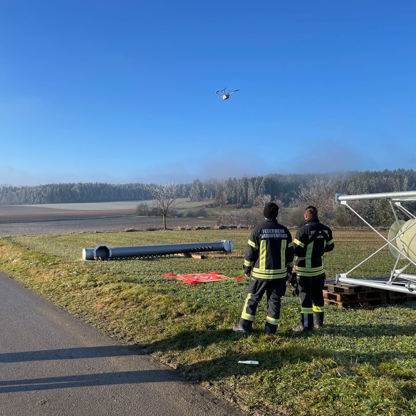 Zwei Feuerwehrleute in Uniform stehen neben einer großen Rohrleitung und einer Metallkonstruktion, mit einem fliegenden Drohnen. Die Szene spielt sich auf einer Grasfläche mit Bäumen und Bergen in der Ferne ab.