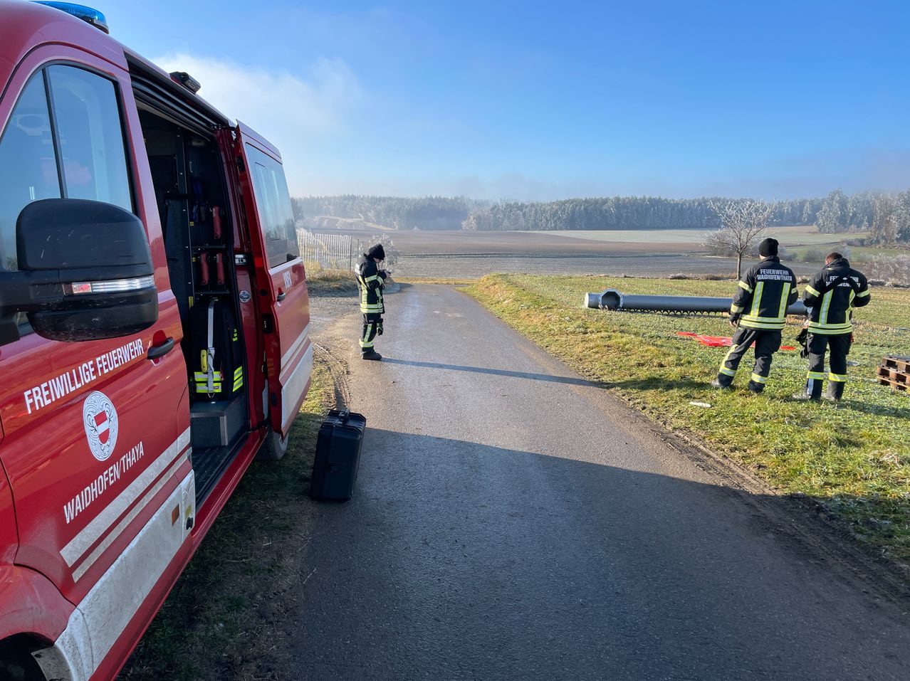 Zwei Feuerwehrleute in Uniform stehen auf einer Straße, einer in der Nähe eines roten Feuerwehrwagens. Ein schwarzer Koffer steht neben dem Wagen. Ein großer Rohr liegt auf dem Gras in der Nähe. Der Himmel ist klar.