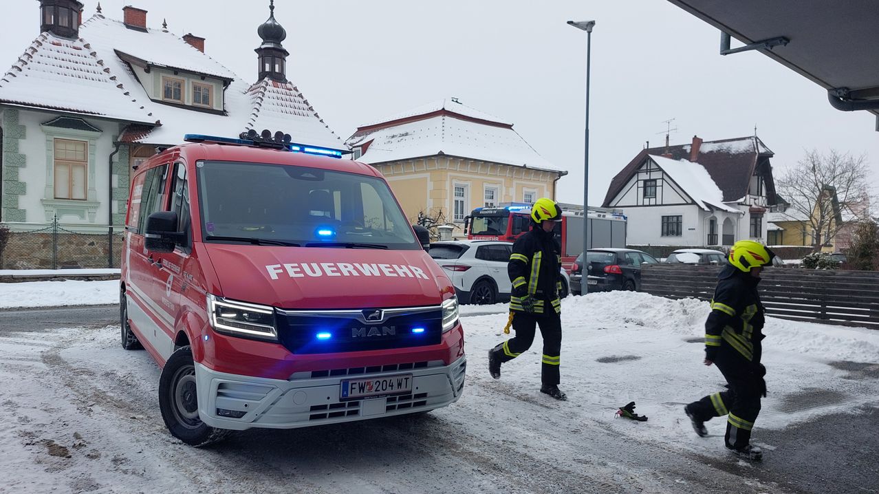 Ein Feuerwehrmann geht auf schneebedecktem Boden neben einem roten Feuerwehrwagen, mit Gebäuden und geparkten Autos im Hintergrund.