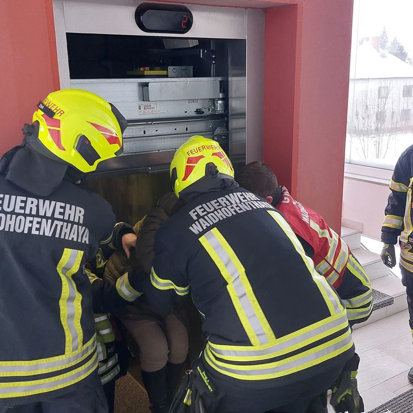 Feuerwehrleute in gelben und schwarzen Uniformen helfen einer Person in einem Aufzug. Der Aufzug ist offen und das Innere ist sichtbar. Die Person trägt eine rote Jacke. Die Szene spielt drinnen mit einer roten Wand.