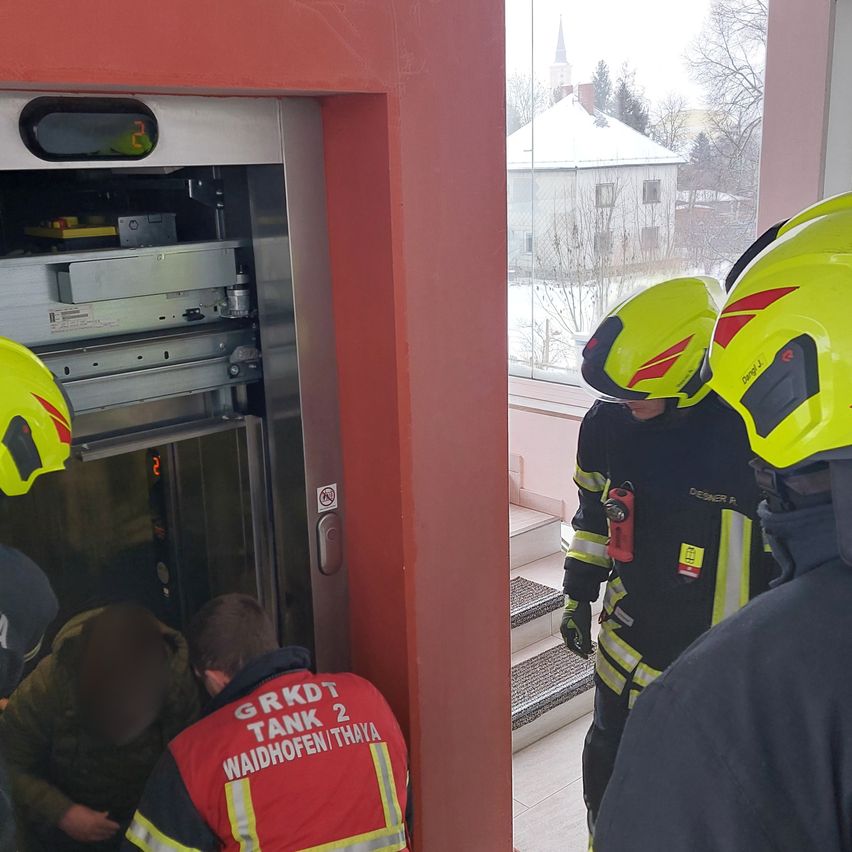 Zwei Feuerwehrleute in gelben Helmen inspizieren den Aufzug, einer kniet im Inneren, während ein anderer draußen steht und zusieht. Der Aufzugschacht ist offen und zeigt Maschinen. Ein Haus mit schneebedecktem Dach ist durch das Fenster sichtbar.