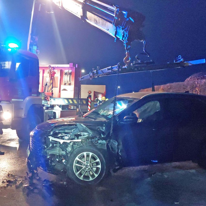 Ein beschädigtes Auto wird nachts von einem Kran angehoben. Notleuchten blinken, und Feuerwehrleute sind in der Nähe. Das Auto ist zerbrochen, mit einer verbogenen Front.