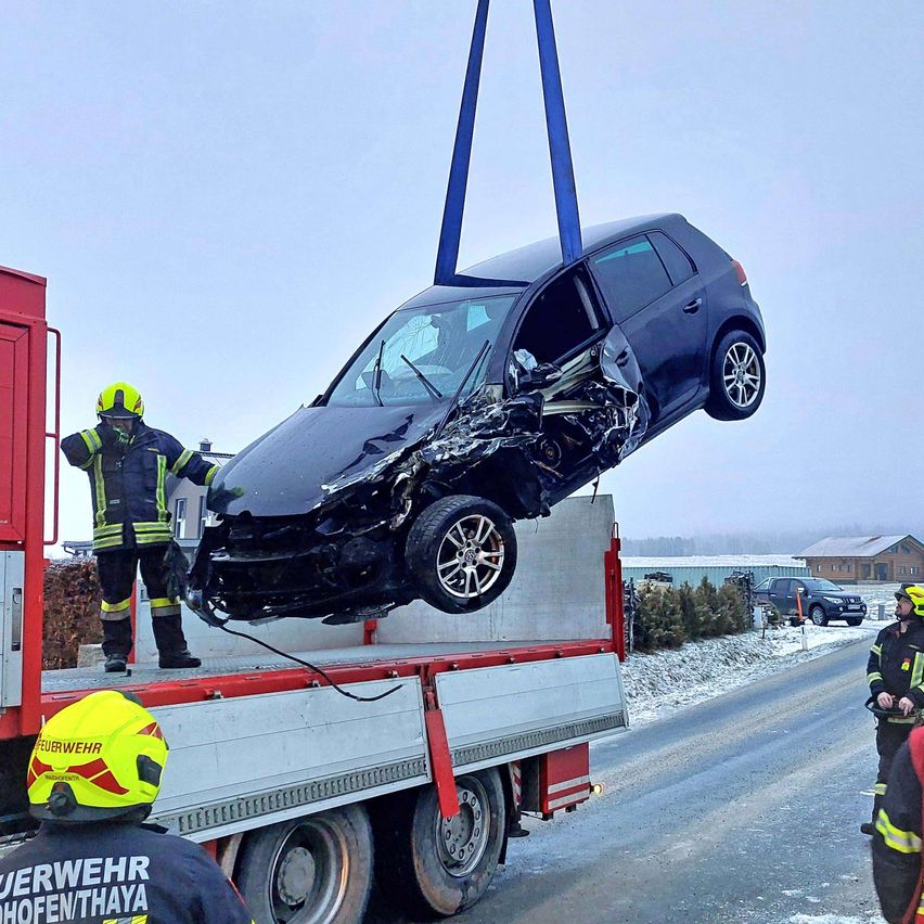 Feuerwehrleute entfernen ein demoliertes schwarzes Auto von einem Lastwagen auf einer verschneiten Straße. Das Fahrzeug hat erhebliche Schäden an der Front.
