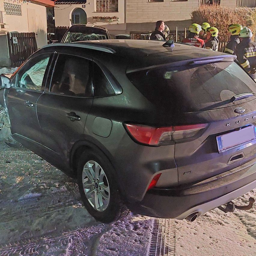 Ein dunkles Auto mit zertrümmerter Rückseite steht auf einer verschneiten Straße, Feuerwehrleute und Schaulustige stehen in der Nähe.