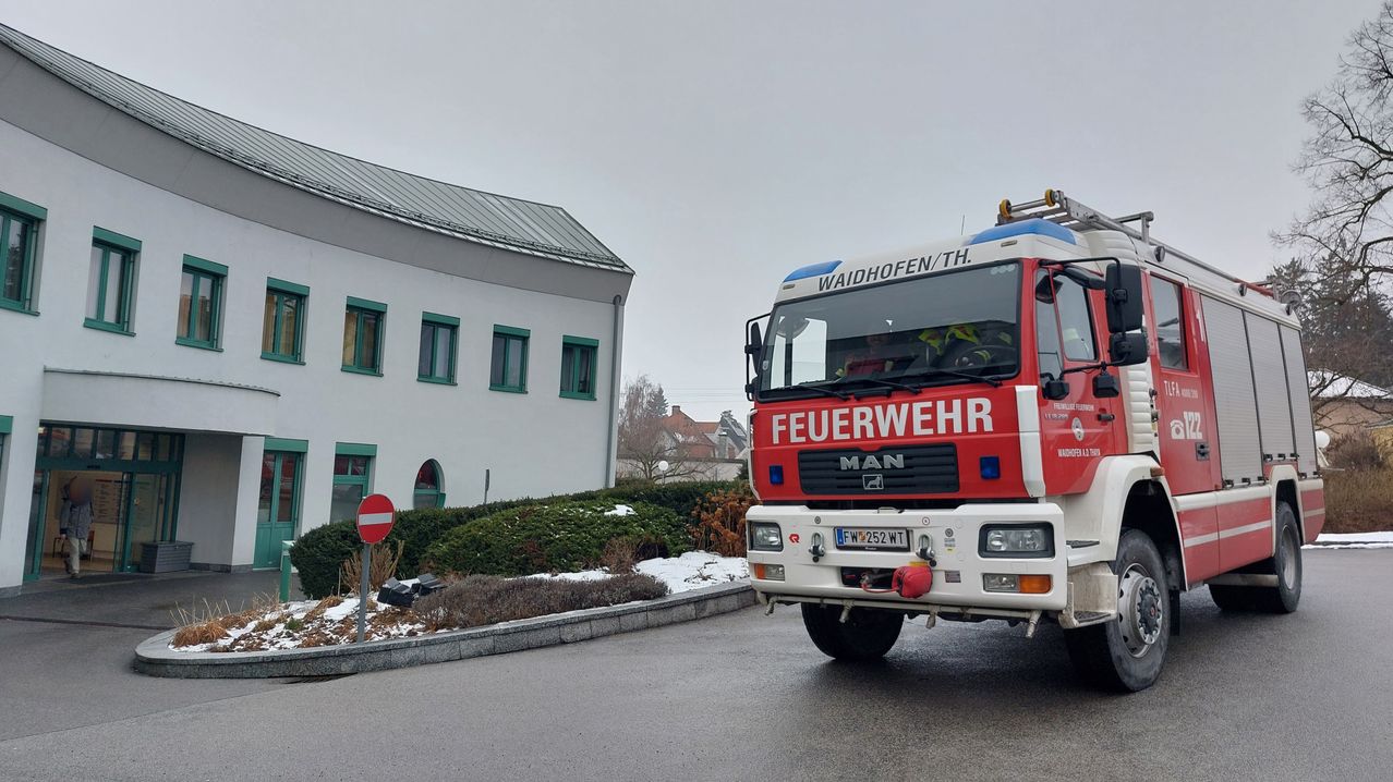 Ein rot-weißer Feuerwehrwagen aus Waidhofen/Thaya fährt eine Straße hinunter, im Hintergrund ein Gebäude mit grünen Fenstern.