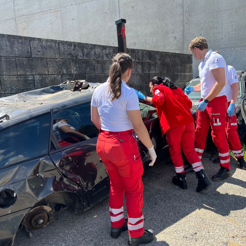 Eine Gruppe von Notfallhelfern in roten Overalls und Handschuhen kümmert sich um eine Person in einem beschädigten Auto. Das Auto ist schwer beschädigt.