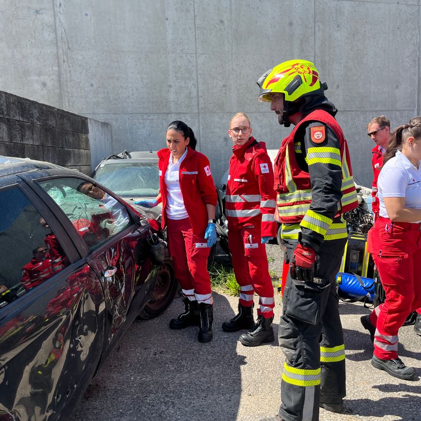 Eine Gruppe von Rettungskräften, darunter ein Feuerwehrmann, kümmert sich um ein beschädigtes Fahrzeug mit einer Frau darin. Eine weitere Frau steht in der Nähe, während andere beobachten.