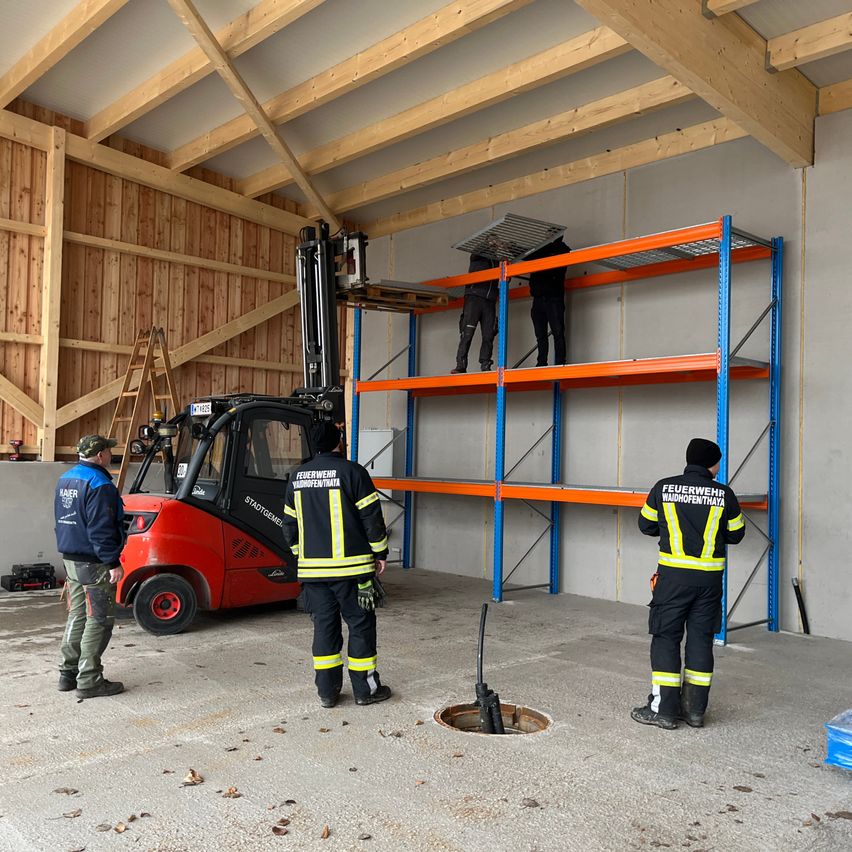 Firefighters are installing shelves in a warehouse using a forklift. Three firefighters are present, one operating the forklift. The shelves are blue and orange.