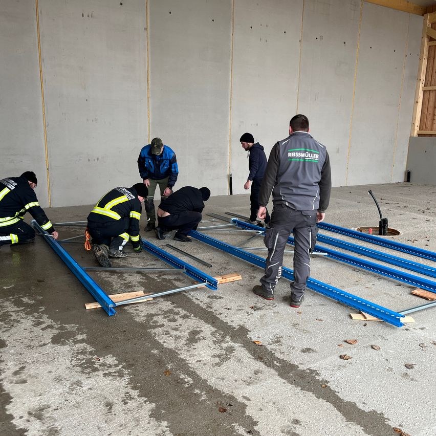 Six workers are assembling a blue metal structure on a concrete floor. They wear jackets and pants, with some wearing helmets. A man in a gray jacket walks among them.