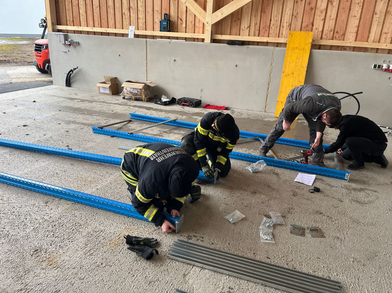 Four firefighters in uniforms are working on the floor of a building, possibly installing a blue metal structure. They are bending down, and one is holding a tool. There are tools, boxes, and other items on the floor. Behind them is a concrete wall.