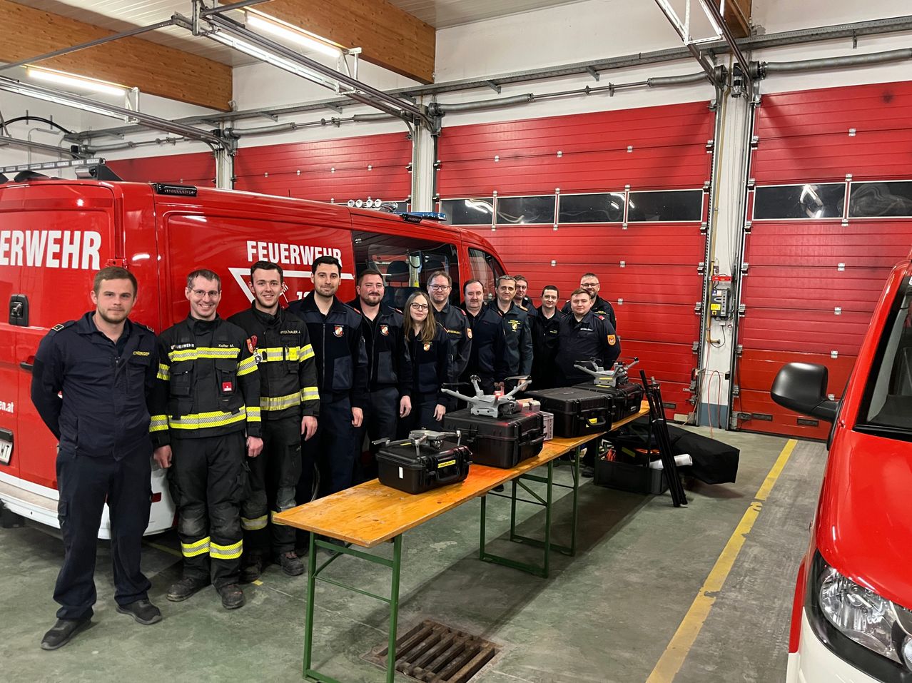 A group of firefighters are standing in a garage with a red fire truck. They are wearing uniforms and posing for a photo. There is a table with several black cases in front of them.