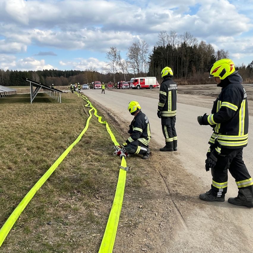 Firefighters are preparing for an emergency response. One firefighter is kneeling, working on a yellow hose, while others stand nearby. In the background, emergency vehicles are parked.