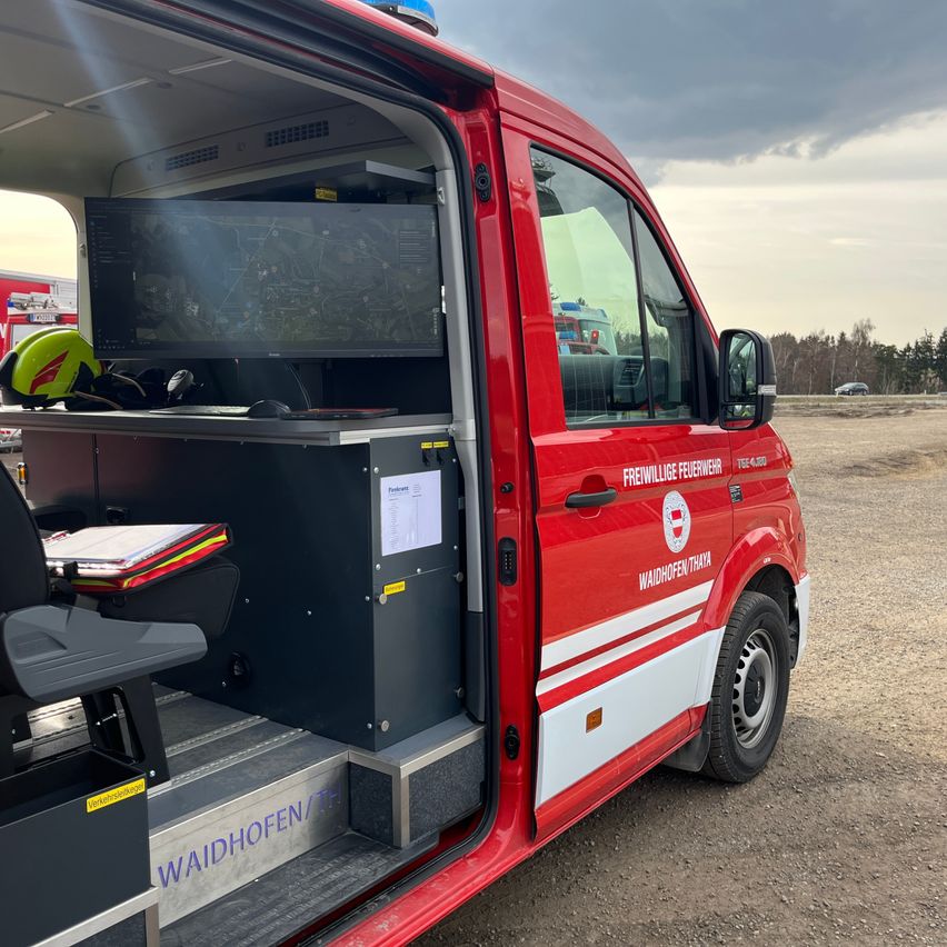 A red fire truck with the door open, showing a monitor, a chair, and various equipment inside. The truck is parked on a gravel surface.