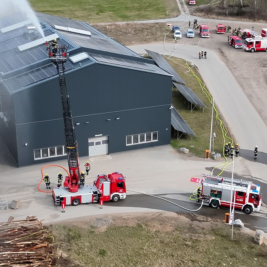 An aerial view of a fire station with multiple fire trucks parked and firefighters in action. The building has solar panels.