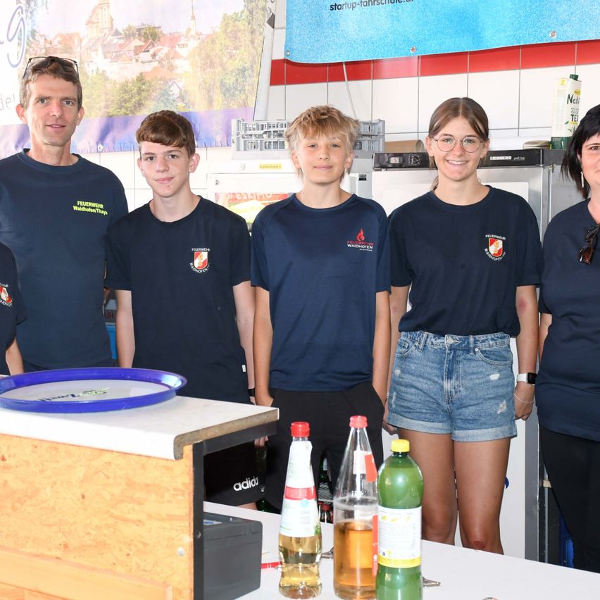 Five individuals, three boys and two girls, are standing together in a kitchen, smiling for a photo. They are wearing matching blue shirts with logos. Behind them, there is a refrigerator and a banner with text. Bottles and a blue plate are on the counter.