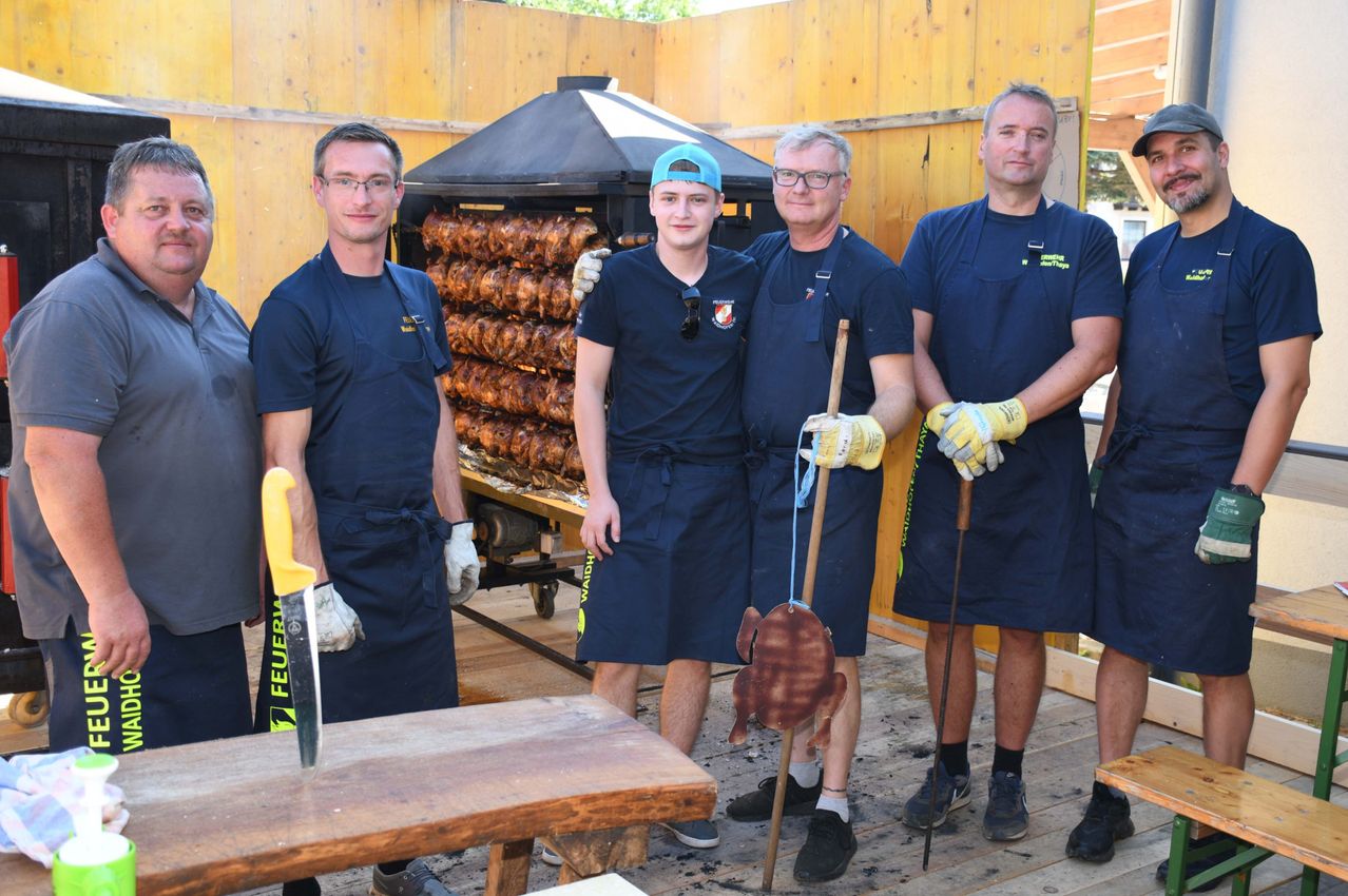 Five men in blue aprons and gloves stand in front of a wooden wall. They are holding a stick with a roasted chicken on it. Behind them is a grill with many roasted chickens.
